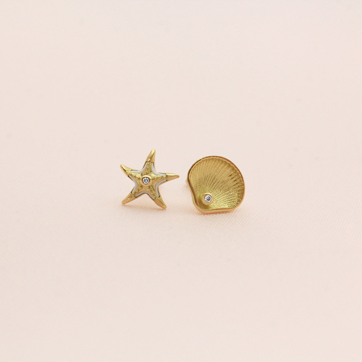 Mismatched gold stud earrings featuring a starfish with white enamel accents and a seashell, each with a small diamond in the center, displayed on a beige  background.