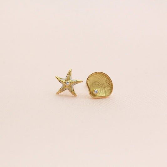 Mismatched gold stud earrings featuring a starfish with white enamel accents and a seashell, each with a small diamond in the center, displayed on a beige  background.
