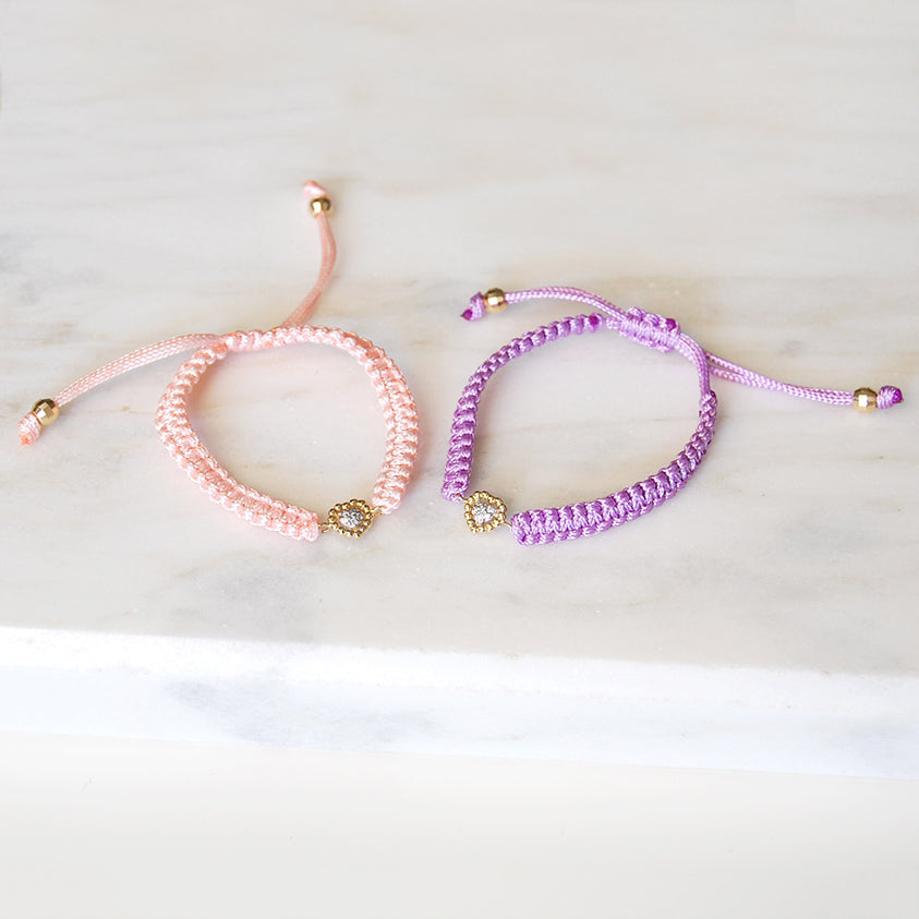 A pair of Sweetest of Hearts diamond charm bracelets in pink and purple cords displayed flat on a marble surface.
