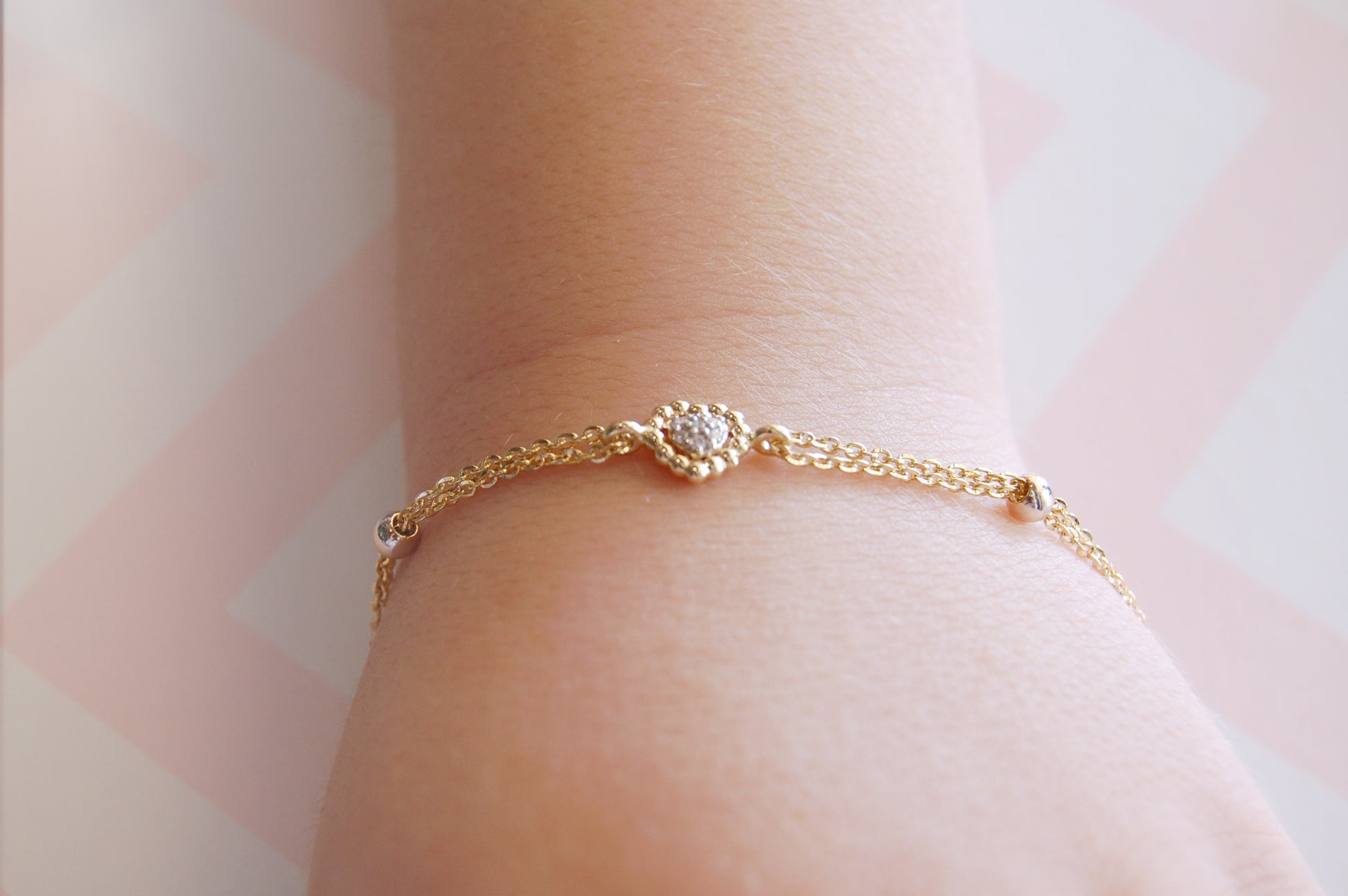 A close-up styling shot of a wrist wearing an adjustable 18K yellow-gold bracelet, featuring a small scalloped-bead heart motif set with a cluster of tiny round diamonds, suspended on a fine double paper-clip chain with sliding gold bead adjusters.