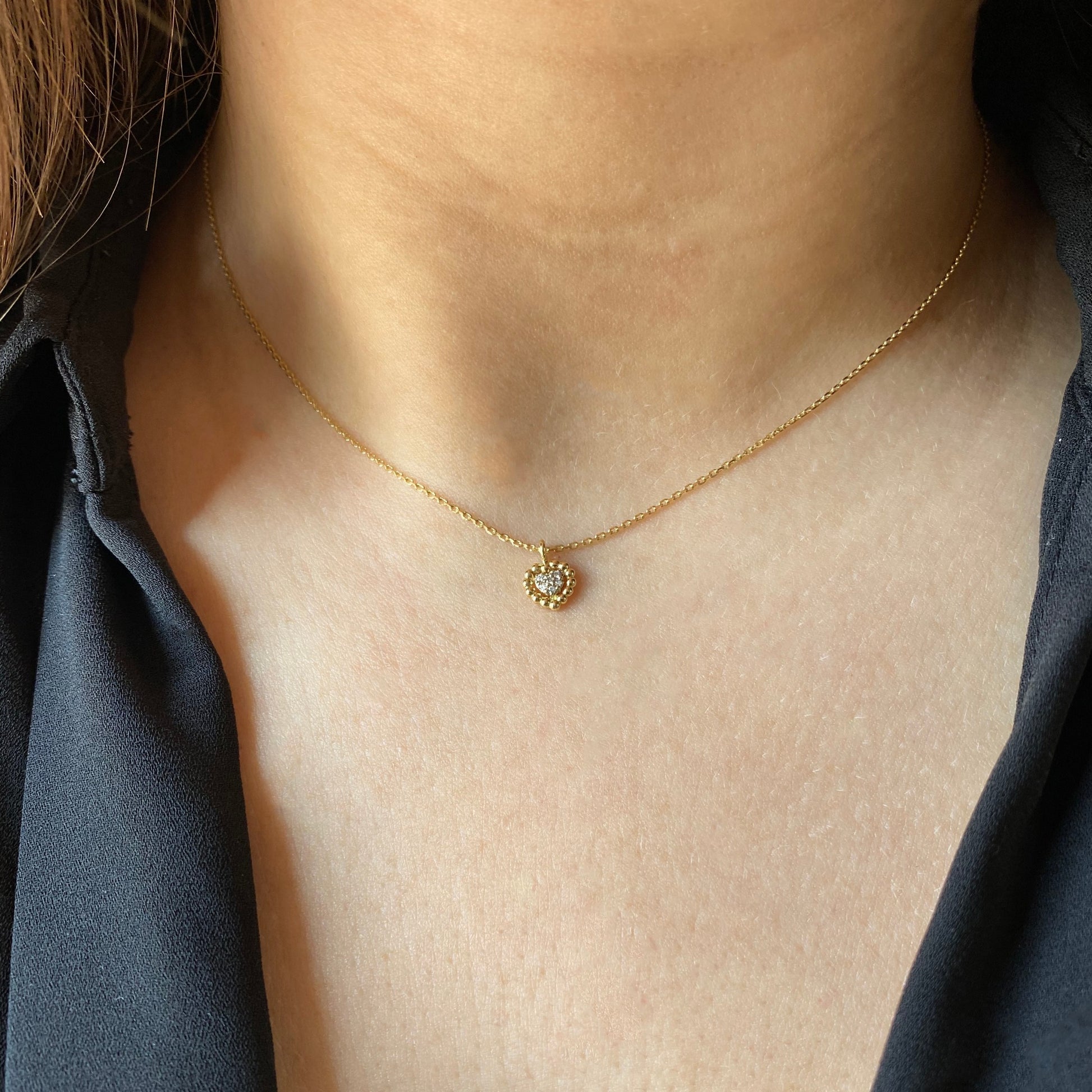 A close-up of a woman’s collarbone wearing a delicate 18K yellow-gold necklace, featuring a small scalloped gold-bead heart pendant set with a cluster of round brilliant-cut diamonds on a fine chain.