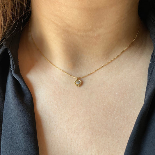 A close-up of a woman’s collarbone wearing a delicate 18K yellow-gold necklace, featuring a small scalloped gold-bead heart pendant set with a cluster of round brilliant-cut diamonds on a fine chain.