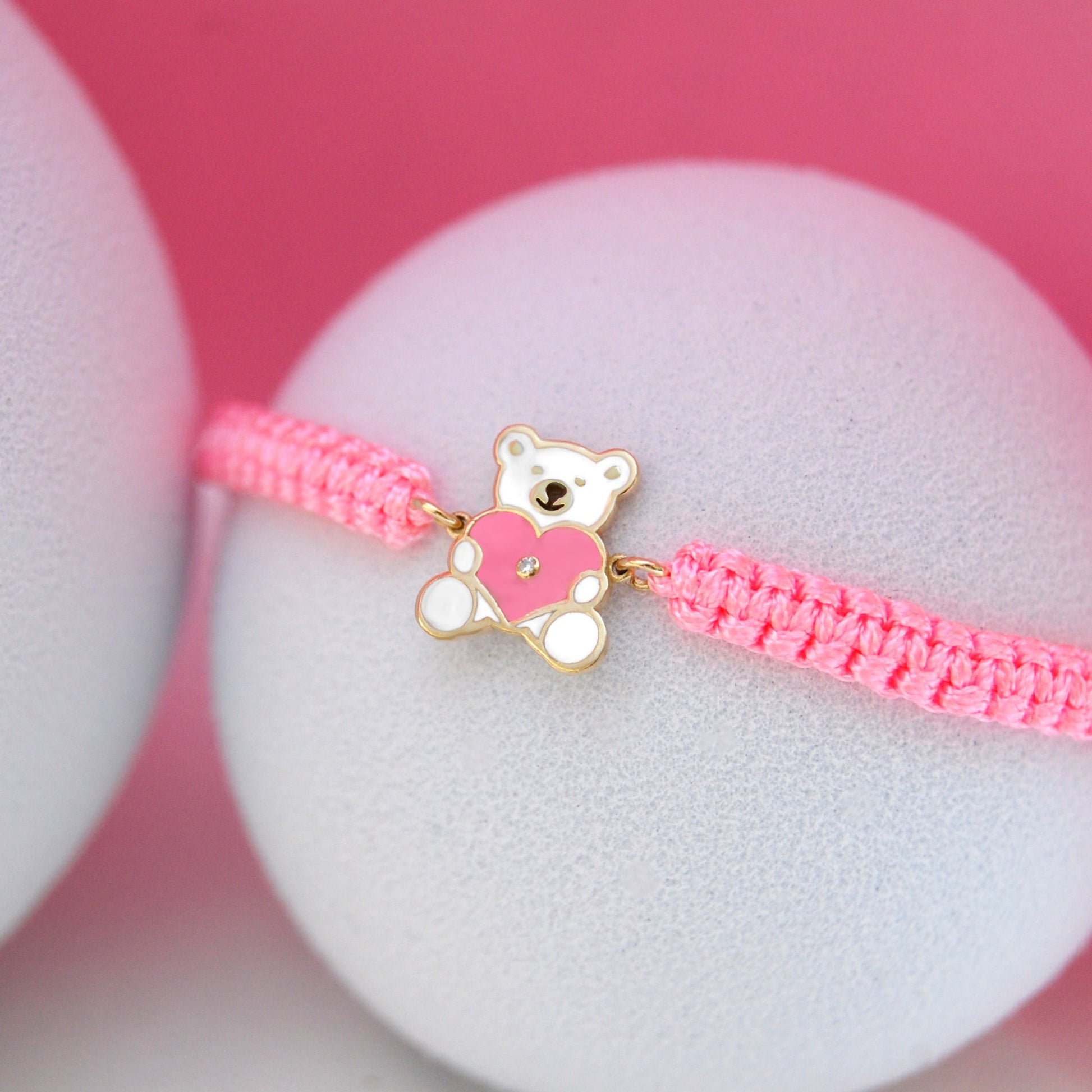 Pink bracelet with a teddy bear charm on a white stand against a pink background
