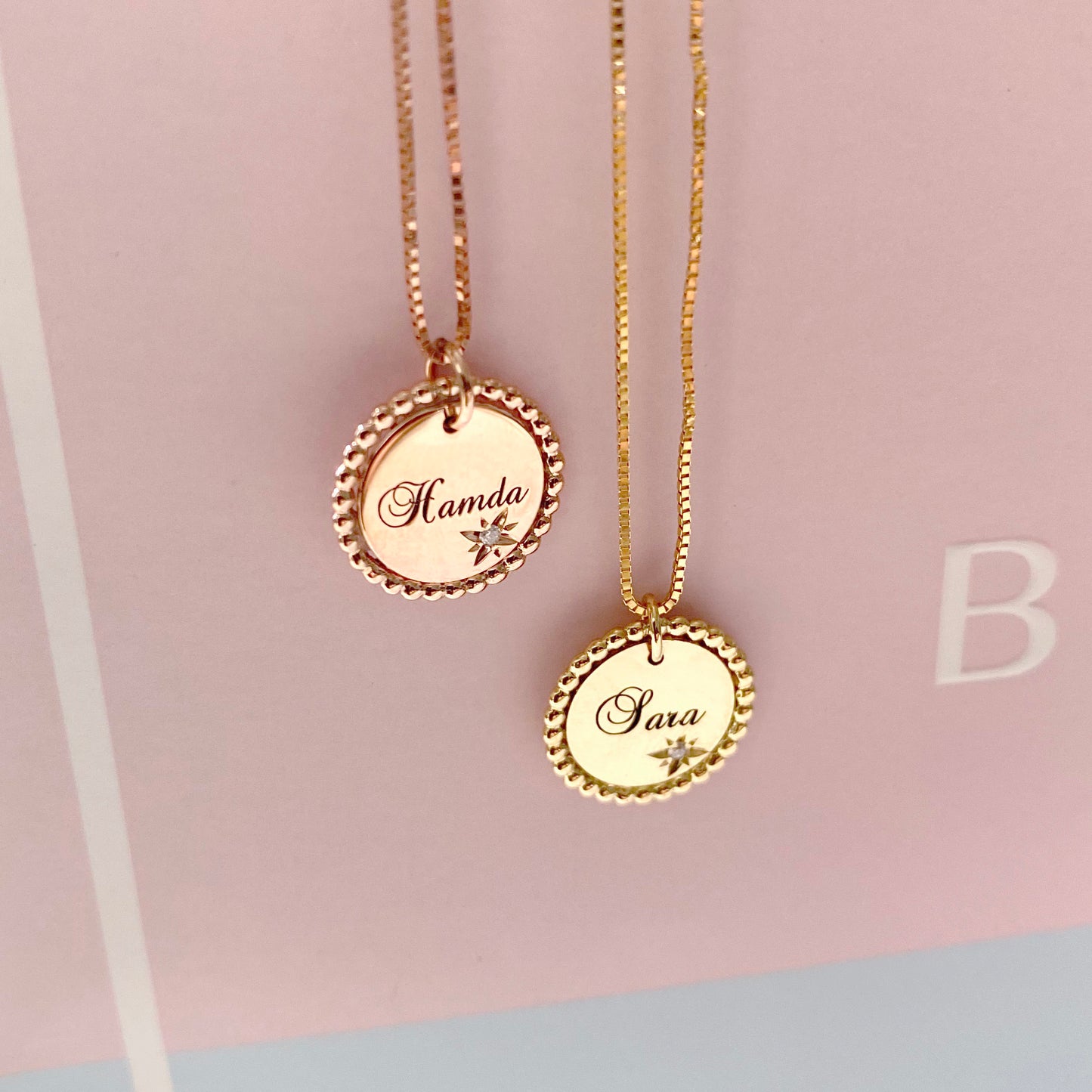 Two 18K yellow-gold scalloped-edge coin pendant necklaces hanging side by side against a blush background—each pendant engraved in script with a name (‘Hamda’ on the left, ‘Sara’ on the right) beside a starburst motif set with a single round diamond, suspended chains.