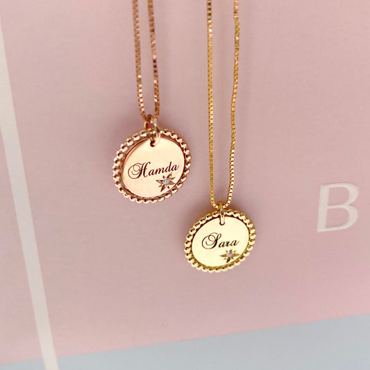 Two 18K yellow-gold scalloped-edge coin pendant necklaces hanging side by side against a blush background—each pendant engraved in script with a name (‘Hamda’ on the left, ‘Sara’ on the right) beside a starburst motif set with a single round diamond, suspended chains.