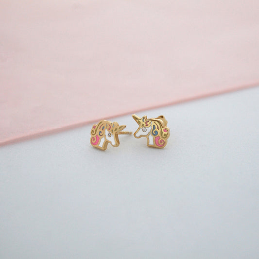 Close-up of a pair of unicorn-shaped stud earrings in gold with multicolor enamel details in pink, white, and blue, and a diamond eye on each unicorn, displayed on a soft pink and white surface.