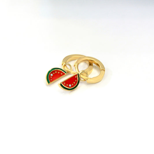 Gold hoop earrings with dangling watermelon slice charms in red and green enamel, each slice accented with a small diamond, placed on a white surface.